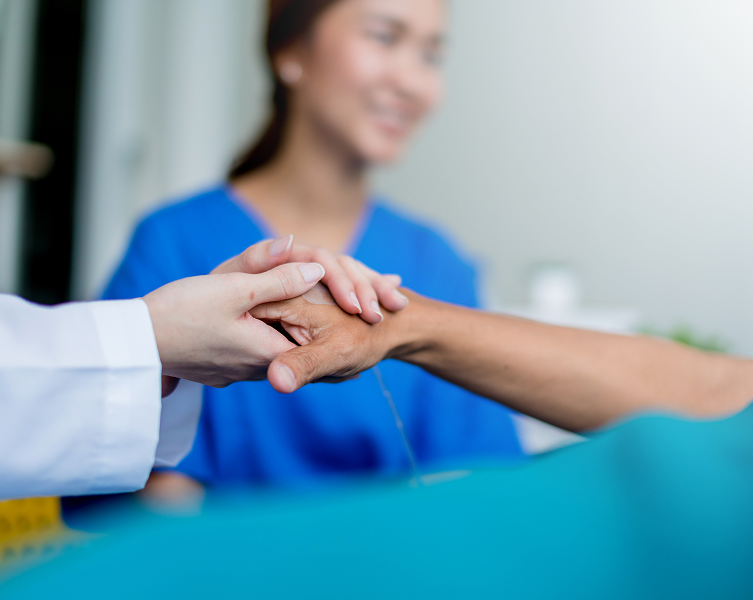 Primo piano di mani che stringono con cura e supporto quelle di un paziente, con un'operatrice sanitaria sorridente sfocata sullo sfondo - Fondo Sanità TLC